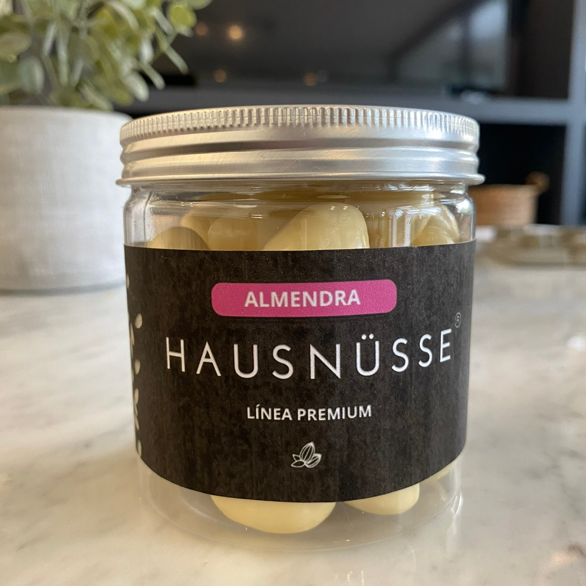 Un frasco de plástico transparente con tapa plateada, con la etiqueta "Hausnusse Almendra Bañada En Chocolate Blanco 250Gr". El tarro contiene almendra bañada en chocolate blanco. Está colocado sobre una superficie de mármol con una planta y un fondo borroso visible.