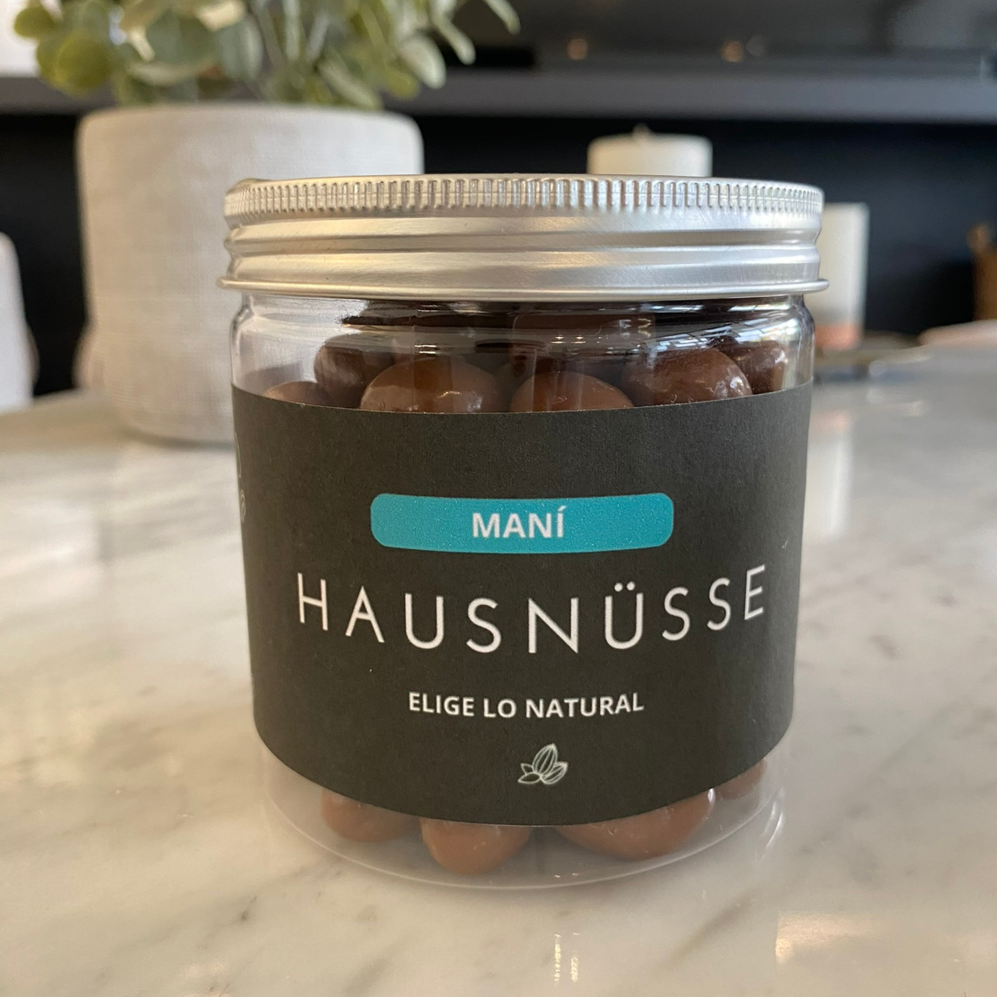 Un frasco de plástico transparente con tapa blanca está lleno de Mani Bañado En Chocolate 250Gr, con la etiqueta "Hausnusse" y "snack saludable" en una etiqueta negra. La etiqueta también dice "Elige lo natural" encima de un pequeño emblema de hoja. El fondo es una superficie de mármol con una planta y objetos borrosos.